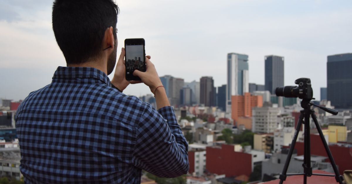Man photographing city skyline with smartphone and camera on tripod from a rooftop.